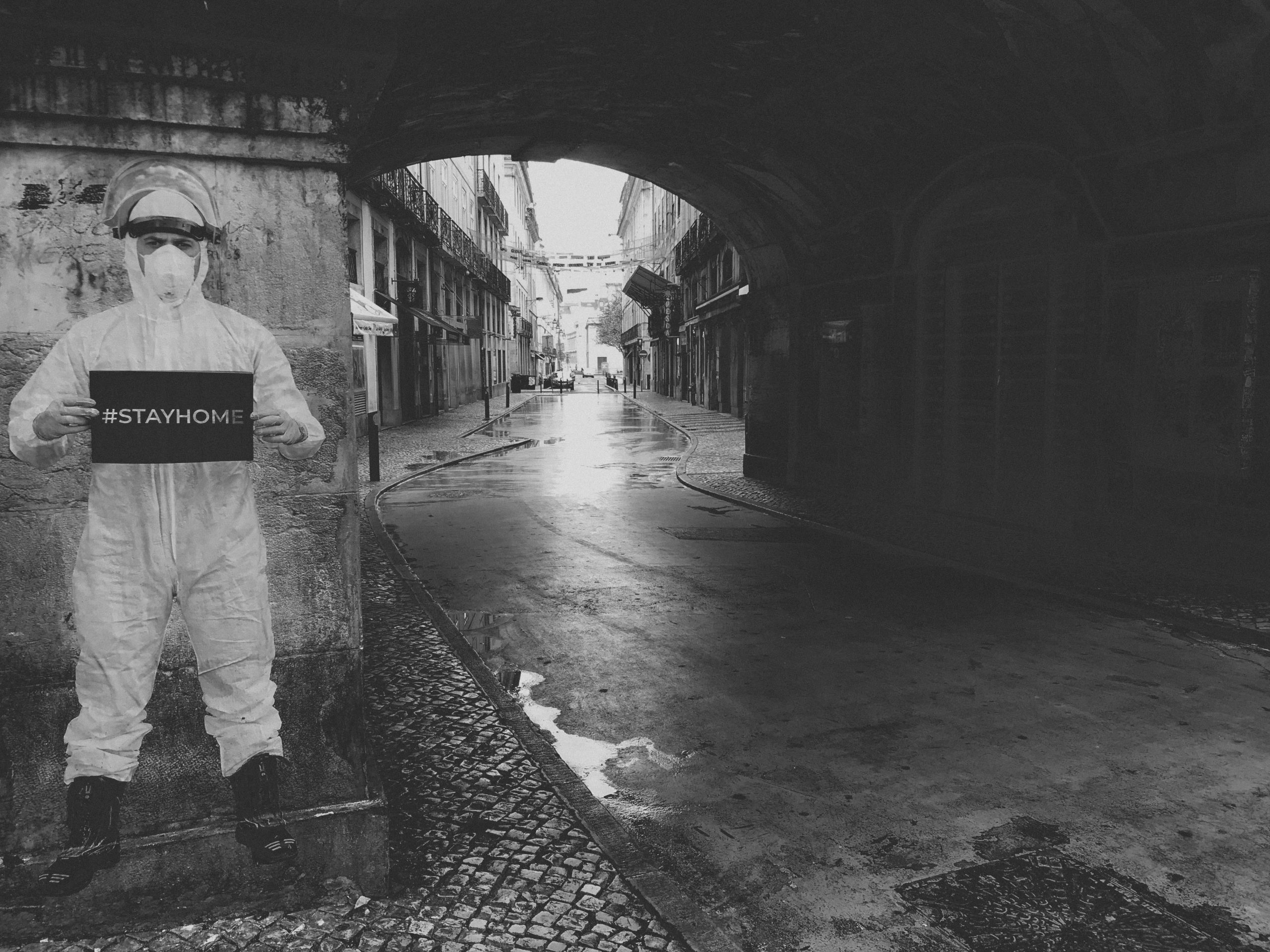 White/black image of a deserted street with an image printed on the wall. The image is a person in protective gear and a face mask, holding a sign that reads "stay home".